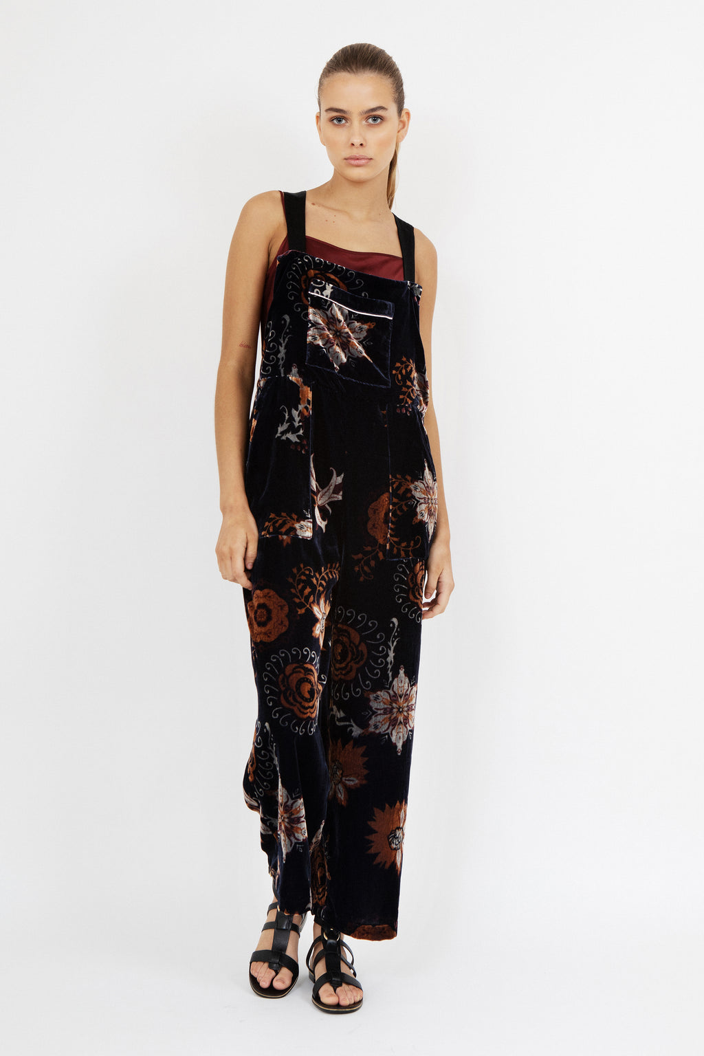 Women’s wide-leg velvet jumpsuit in deep navy with burnt orange and beige floral print, featuring square neckline, front pocket detail, and adjustable straps – model wearing black sandals