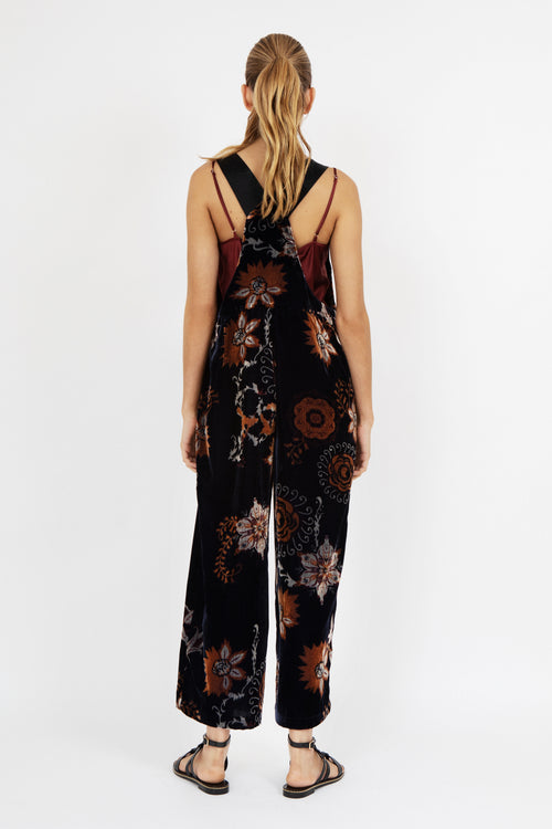 Women’s wide-leg velvet jumpsuit in deep navy with burnt orange and beige floral print, featuring square neckline, front pocket detail, and adjustable straps – model wearing black sandals